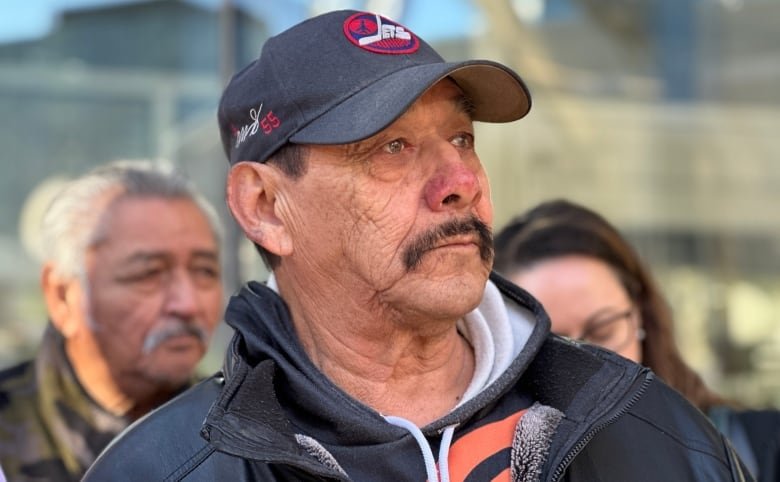 A teary-eyed man wearing a baseball cap and hooded sweatshirt is seen in a close crop, showing his head and shoulders.