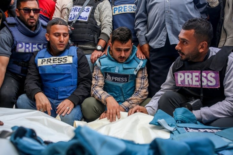 Four men in press vests kneel in front of bodies covered in white and laid out on the ground.