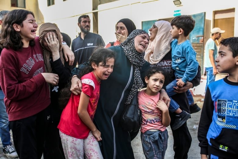 A group of women, children and men stand in the streets crying and screaming as they look toward something off camera.