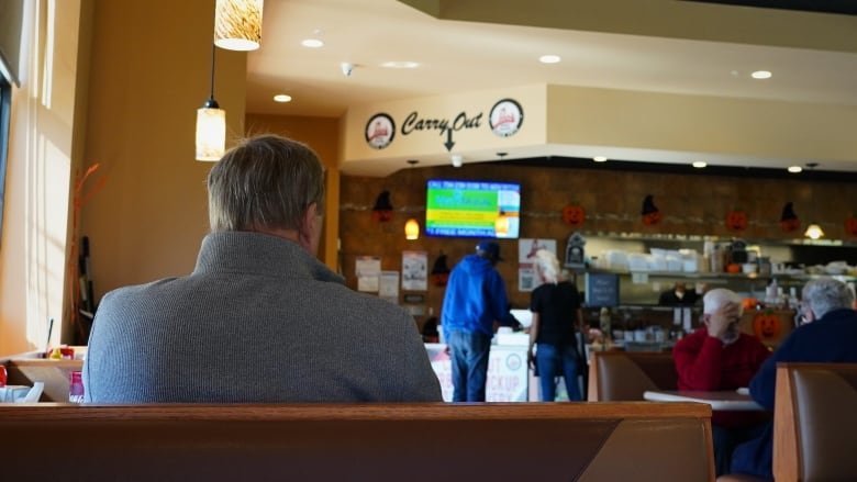 A grey-haired man in a grey sweater is seen inside a booth at an American diner. Halloween decorations are in the background.