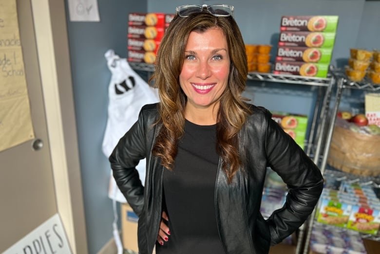 A smiling woman wearing all black stands in front of wire shelving holding crackers, fruit cups, barrels of apples, granola bars and cereal bowls.