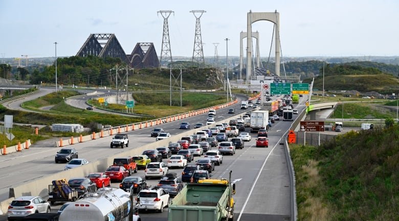 A large amount of cars and trucks are backed up together in a line in the right lanes of a highway. There are only a few cars on the left lanes. In the distance, the tall spokes of a bridge are visible, as are some electrical transformers and the triangles of yet another bridge off to the left.