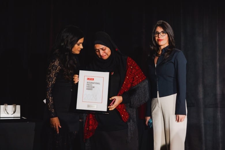 A crying woman in a hijab holds stands flanked by two other woman as she holds up a framed piece of paper with the words: " International Press Freedom Award."