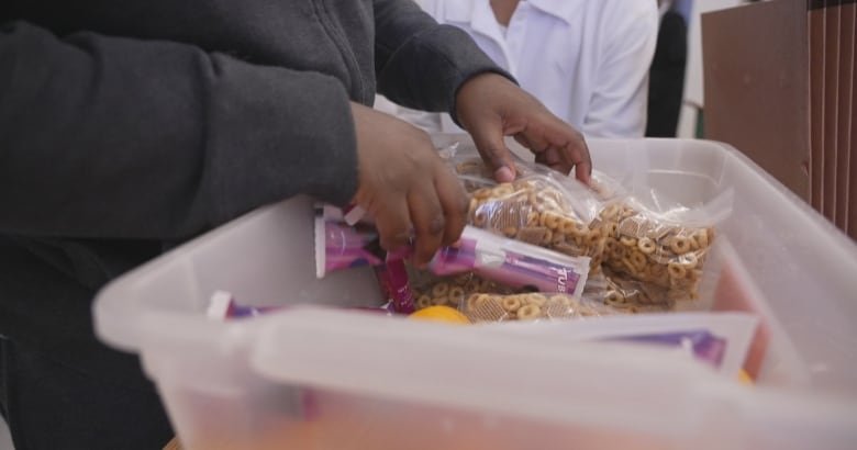 A young person's hands are shown rifling through a snack bin filled with ziplocks of cheerios, oranges and tubes of yogurt.