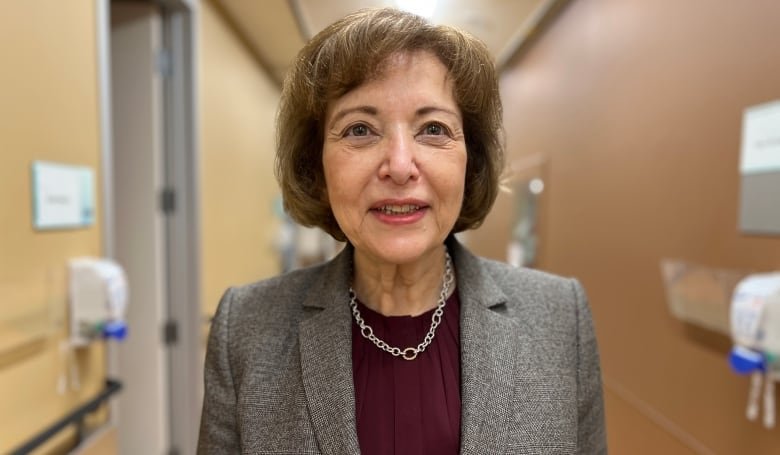 A woman with short brown hair is wearing a a tweed jacket and a purple blouse.