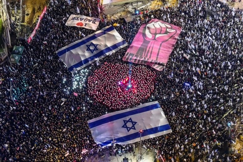 Aerial view of thousands of people on the street at night, holding lights and large Israeli flags.