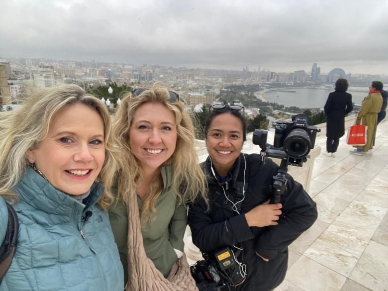 Three women, one with a camera, at a high view point, with a smoggy city and lake int he background.